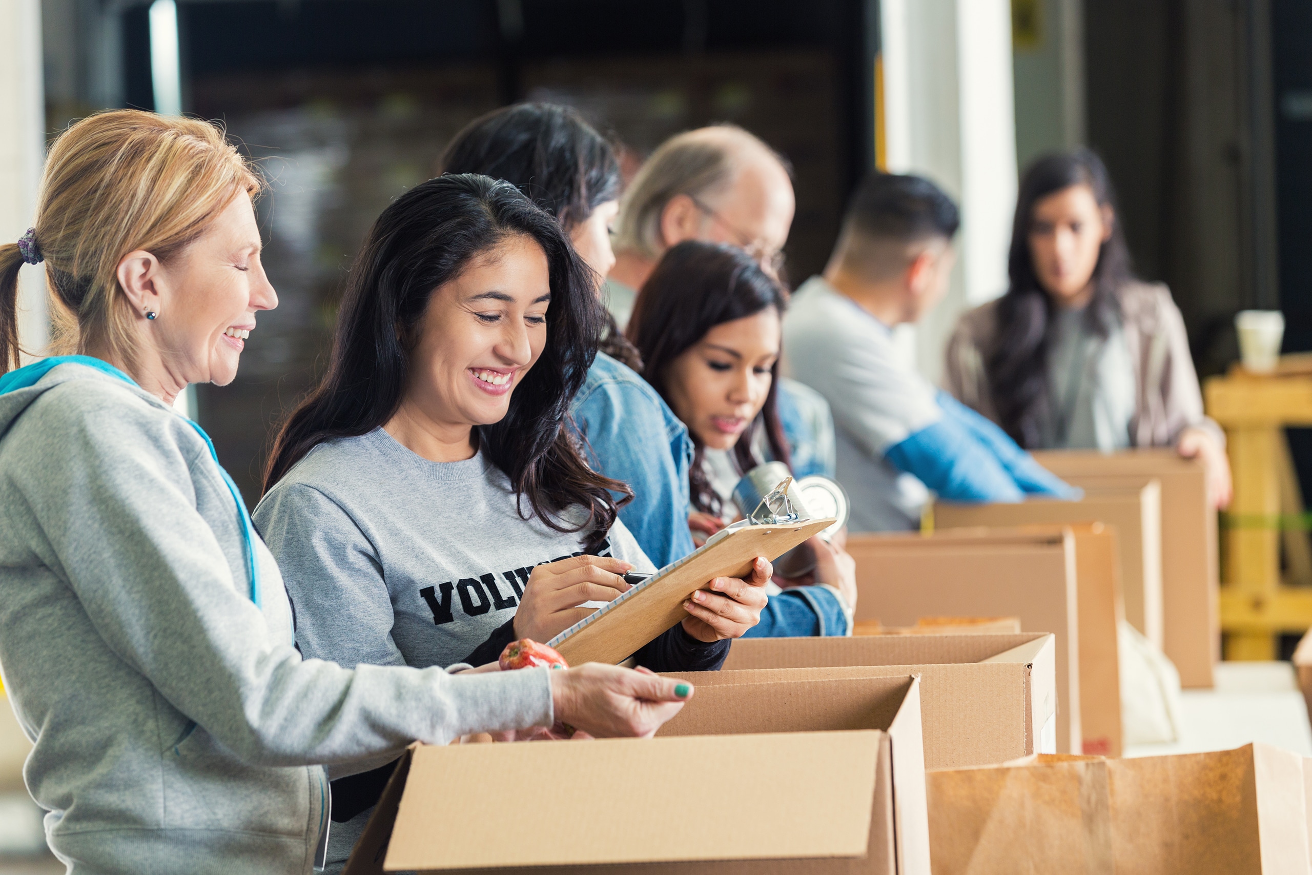 Mature adult Caucasian woman is standing with mid adult Hispanic woman as they pack cardboard boxees full of donated food in charity food bank. Other volunteers are lined up behind them, also sorting donated groceries into boxes. Hispanic woman is writing on checklist on clipboard.